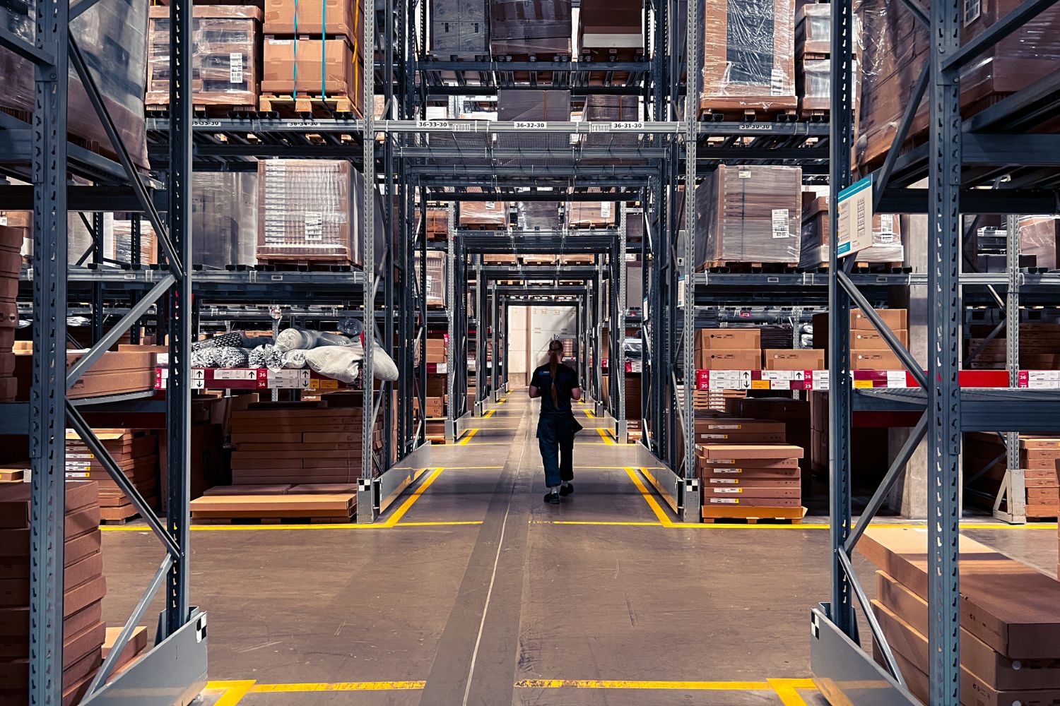  Image of a warehouse with many boxes and a worker walking down the aisle
