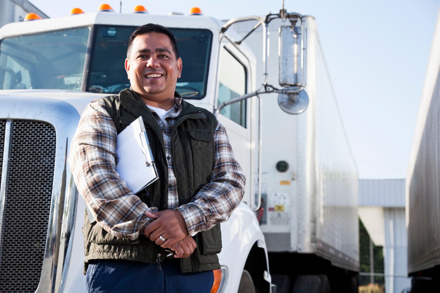 Image of a man smiling while holding a clipboard in front of a transportation truck